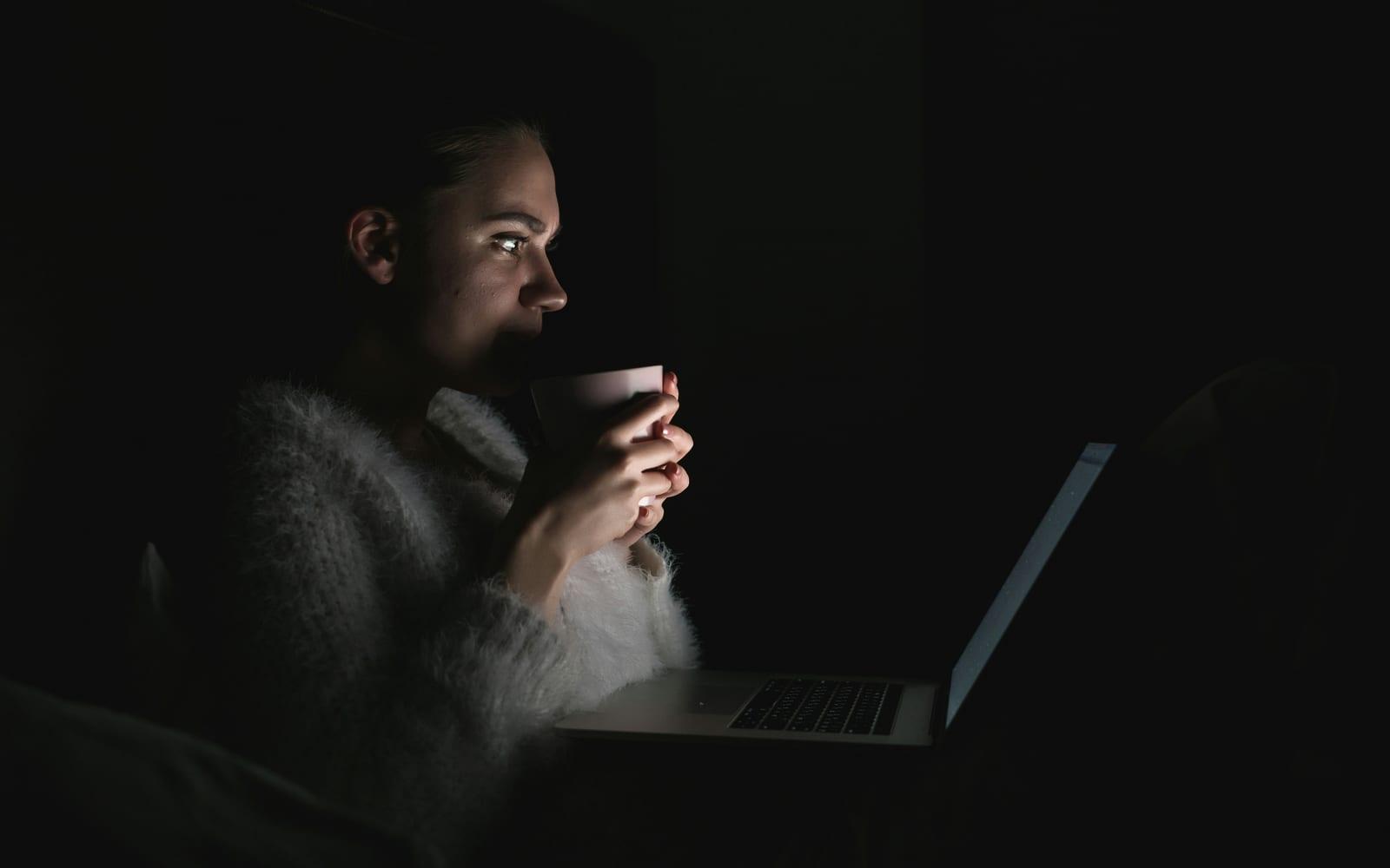A woman sits awake in the dark, lit only by her laptop screen, holding a mug